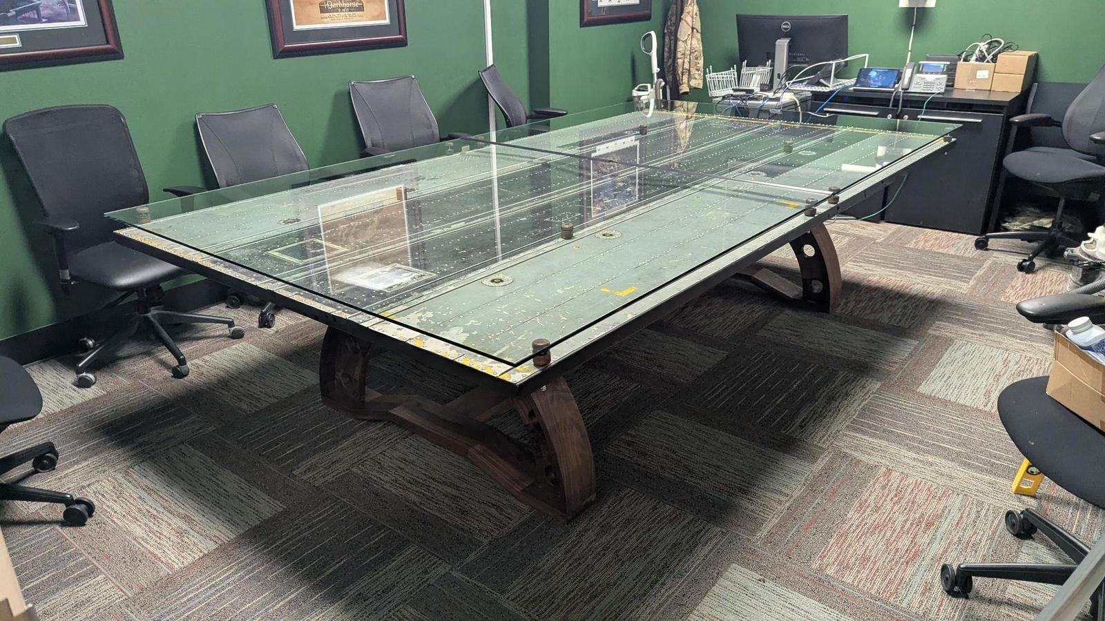 Large glass-topped conference table with riveted metal panels beneath the glass, installed in a green-walled conference room
