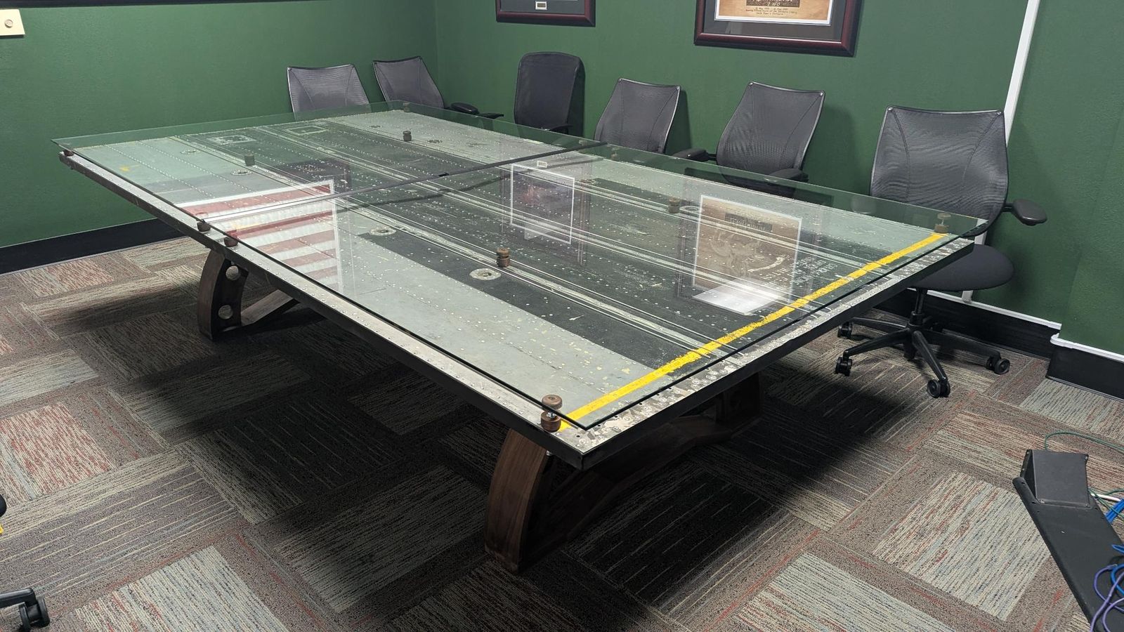 Corner view of the conference table showing riveted panels and a yellow edge stripe beneath the glass top